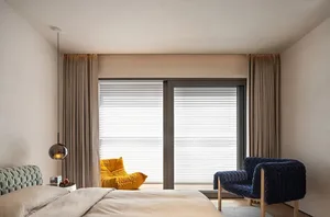 A modern bedroom featuring a beige fabric bed with a yellow and blue upholstered chair in front of a large window with blinds