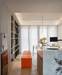 A modern residential space featuring white marble countertops, orange stools, and a bookshelf filled with books.