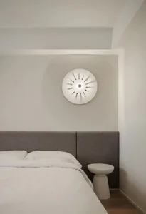A minimalist bedroom featuring a white bed with a gray upholstered headboard against a light-colored wall, illuminated by a u