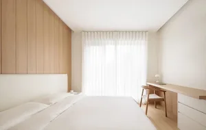 Modern bedroom with white fabric bedding, wooden headboard, and large window with sheer curtains.