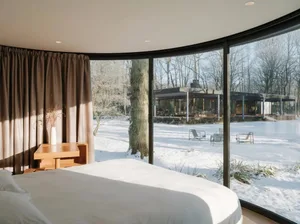 A minimalist bedroom with a white bed, glass windows overlooking a snowy landscape, and modern furnishings.