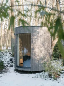 Modern outdoor sauna constructed from wood with a large glass window, situated in a snowy forest.