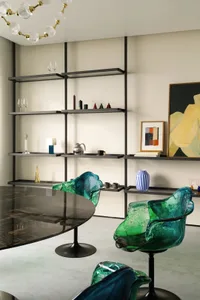 Modern dining area featuring sleek glass table, green and blue sculptural chairs, and open shelving.