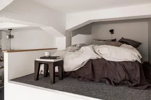 A minimalist bedroom with a bed covered in beige and brown bedding, a wooden bedside table, and concrete walls.