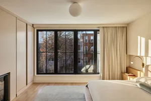 A minimalist bedroom featuring a large bed with white linens, a wooden headboard, and a large window with beige curtains and