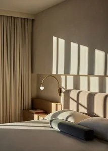 A minimalist bedroom with a bed, pillows, and a wall-mounted light fixture casting shadows on the wall.