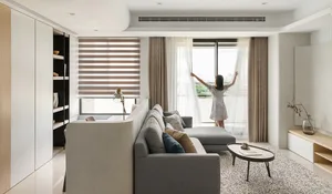 Person adjusting beige curtains in a minimalist living room with a gray sectional sofa and a round coffee table.