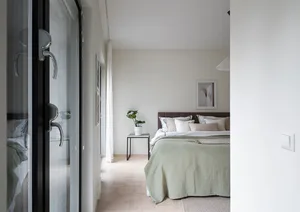 A minimalist bedroom featuring a bed with a muted green bedspread and a headboard, accompanied by neutral-toned bedding and a