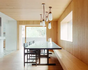 A modern, minimalist dining room featuring a wood table and chairs, with a large window and pendant lights.