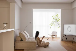 A minimalist living room with a beige fabric sofa, a tree by the window, and a woman sitting on the floor with a drink.