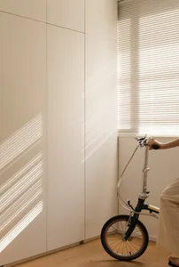 A modern minimalist interior with a bicycle parked next to a window with blinds, casting lines of light into the room.