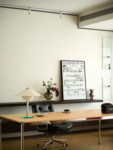 A minimalist office desk with a wooden surface, a black swivel chair, a lamp, a vase with flowers, and a framed architectural