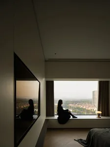 A person sitting on a window sill in a modern interior with glass walls, wooden flooring, and textile furnishings.