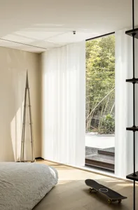 A minimalist bedroom with white curtains and a view of nature, featuring wood flooring and a skateboard.