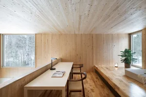 A modern home office with wooden walls and floors, featuring a long table and chairs, surrounded by large windows overlooking