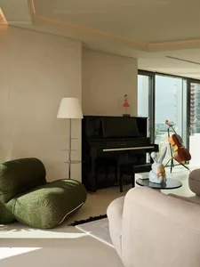 A modern living room with a green beanbag chair, beige sofa, black upright piano, and a cello in the corner.
