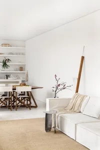 A minimalist living room with a white sofa, wooden floor, and white walls, featuring a wooden stand and a plant.