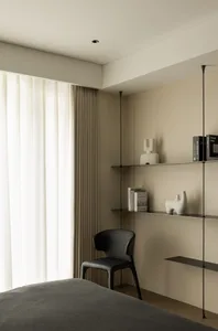 A modern minimalist bedroom with a black chair, floating shelves, and neutral tones on the walls and curtains.