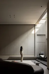 A minimalist living room with concrete walls, a large window, and a person standing in the center.