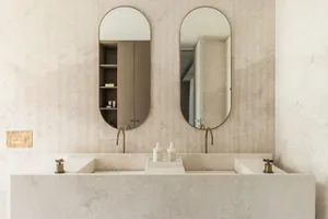 Modern minimalist bathroom with marble countertops and sinks, featuring brass faucets and a light-colored textured wall.