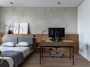 A modern minimalist bedroom featuring a wooden desk with a computer monitor, a bed with white and gray bedding, and a concret