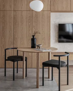 A photograph of a minimalist dining room featuring a round wooden table, two black chairs, and a vase with dried branches.