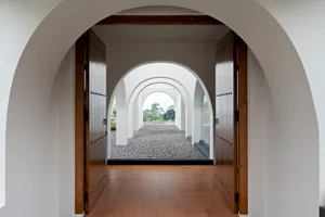 A minimalist entrance featuring a row of archways with concrete walls and wooden doors, leading to a gravel pathway.