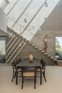 A modern dining room with a concrete wall, a table with chairs, and a person walking on stairs in the background.