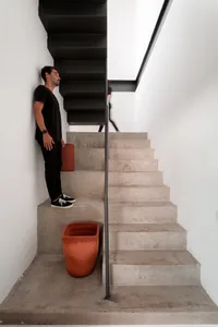 A man standing on a modern concrete staircase with a black railing, wearing a black shirt and sneakers, carrying a red cylind