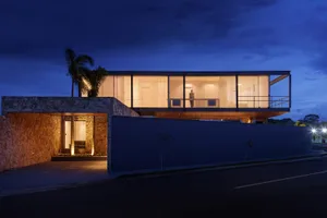 A modern house with glass walls, steel railings, and concrete features, illuminated at dusk.