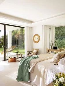 A minimalist bedroom featuring textured fabric bedding, white walls, and large windows overlooking a garden.