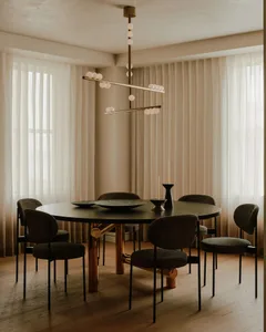 A modern dining room featuring a round wooden table with brass legs and chairs, set against sheer curtains.