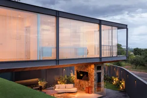 A modern house with a glass exterior featuring vertical blinds and a stone patio.