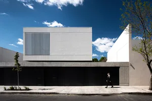 A minimalist building made of concrete with a large window and a person walking on the sidewalk.