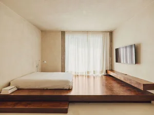 Minimalist bedroom featuring a bed on a raised wooden platform with concrete accents and a window with white curtains.