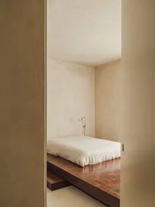 Minimalist bedroom featuring a white bed on a wooden platform and a wall-mounted lamp.