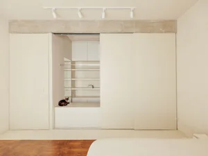 A minimalist bedroom with concrete flooring and walls, featuring a bed and a sliding door leading to a closet.