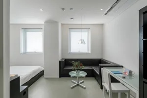 A minimalistic modern studio apartment with metallic accents and a fabric sofa, featuring a glass table and white walls.