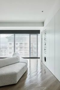 A minimalist living room with a gray sofa and wooden floor, large windows covering the far wall, and white storage units on t
