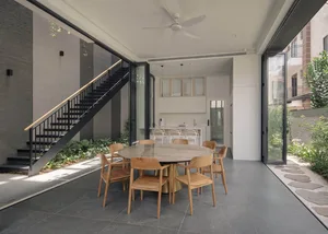 A modern residential dining room featuring a round marble table surrounded by wooden chairs, with a staircase and greenery in