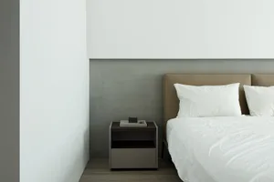 A minimalist bedroom featuring a concrete headboard and a white bed with a book and stone sculpture on a nightstand.