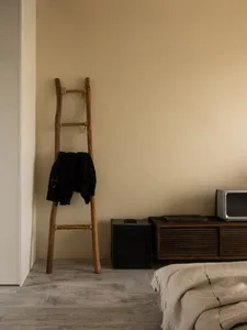 A minimalist bedroom featuring a wooden ladder with a blanket on it, against a beige wall, with rustic wooden furniture and a