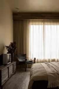 A minimalist bedroom featuring a bed with neutral-toned textiles, a wooden armchair, and a vintage television on a rustic cab