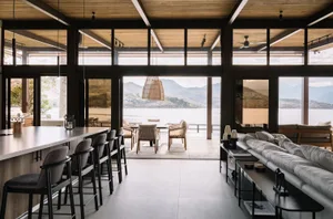 Modern living room with glass walls overlooking a beautiful lake and mountains, featuring sleek furniture and large windows f