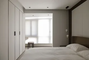 A minimalist bedroom with white and beige neutral colors, featuring a bed, a window with blinds, and a small chair in the cor