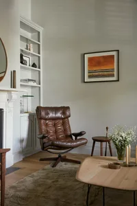A minimalist living room featuring a brown leather reclining chair, a wooden coffee table, bookshelf, and framed artwork