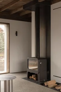 Modern black metal hearth with wood logs and a glass front, set against a minimalist white wall and wooden ceiling beams.