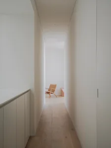 A narrow hallway with white walls and wooden flooring, featuring a single wooden chair in the distance.