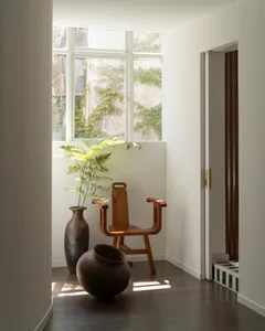 A minimalist indoor space featuring a wooden chair, vases, and a plant, with a white wall and hardwood floor.