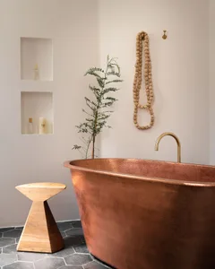 A modern bronze bathtub in a minimalist bathroom with a small wooden stool and green plant.